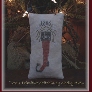 May include: A beige fabric stocking with a red boot and the year 1806 embroidered on it. The stocking is hanging from a branch of a Christmas tree with white lights.