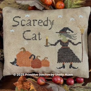 Op de afbeelding: Een gestikt Halloween-kussen met de woorden "Scaredy Cat". Het ontwerp toont een heks met een kaars en een bezem, met pompoenen en een zwarte kat. Het kussen is omgeven door herfstbladeren.