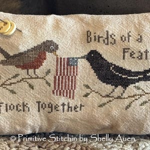 Può includere: Un cuscino beige con un ricamo a punto croce di due uccelli su rami che tengono una piccola bandiera americana. Il testo "Birds of a Feather Flock Together" è ricamato sotto gli uccelli. Il cuscino è firmato "Primitive Stitchn by Shelly Auen".