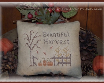 May include: A beige, square decorative pillow with a cross-stitch design. The design features a tree, pumpkins, and the words "Bountiful Harvest". The pillow is surrounded by pine cones and fall foliage.