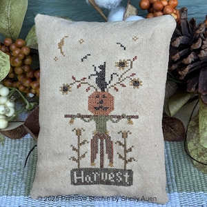 Op de afbeelding: Een beige, rechthoekig decoratief kussen met een kruissteekontwerp met een pompoen, zwarte kat en het woord "HARVEST". Het ontwerp bevat zonnebloemen, een maan en vogels. Het kussen heeft een herfst- of Halloween-thema.
