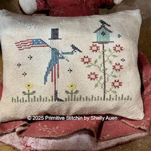 May include: A decorative pillow with a cross-stitch design featuring an Uncle Sam figure holding an American flag. A birdhouse and flowers are also depicted. The pillow is on a red fabric with a floral pattern.