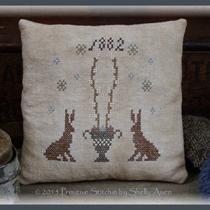 May include: A beige cushion with a cross-stitch design featuring two brown rabbits, a vase with white flowers, and the text "1882". The cushion is surrounded by a wooden frame.