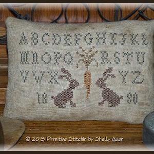 May include: A beige cushion with a cross-stitch design featuring the alphabet, two bunnies, and a carrot. The text "2013 Primitive Stitchin by Shelly Aven" is at the bottom of the cushion.