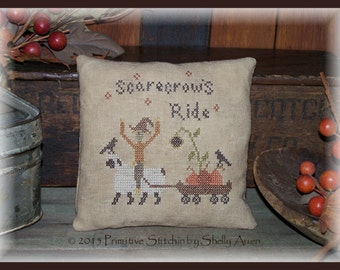 May include: A cross-stitch pillow featuring a scarecrow riding a white animal pulling a wagon full of pumpkins. The pillow is beige with brown stitching and the text "Scarecrow's Ride" in brown lettering.