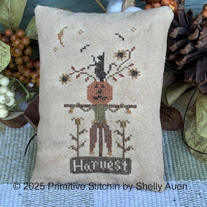 May include: A beige, rectangular decorative pillow with a cross-stitch design featuring a pumpkin-headed scarecrow, a black cat, and the word "Harvest." The design includes a crescent moon and birds. The pillow is surrounded by fall foliage.