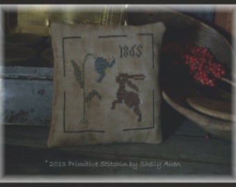 May include: A beige square pillow with a cross-stitched design featuring a rabbit, a flower, and the number "1865". The pillow has a dark border and is set against a rustic background. The text "2013 Primitive Stitchin by Shaly Auen" is at the bottom.