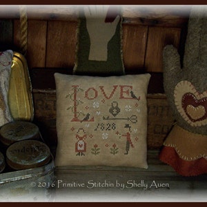 May include: A beige pillow with the word "LOVE" embroidered in red and brown. The pillow also features a small key, a padlock, and two birds. The text "1828" is also embroidered on the pillow. The pillow is sitting on a wooden surface.