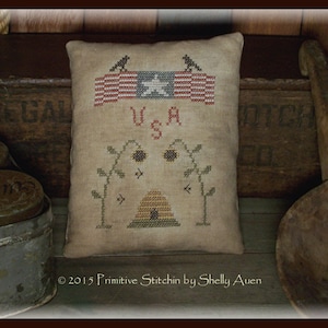 May include: A beige pillow with a cross-stitch design featuring a beehive, sunflowers, and the words "U.S.A." above a red, white, and blue American flag.