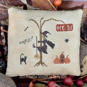 Può includere: Un cuscino quadrato beige ricamato a punto croce con una scena di Halloween. Il disegno include una strega, un gatto nero, zucche e la scritta "Oct. 31". Anche la scritta "oops!" è ricamata sul cuscino. Le parole "2025 Primitive Stitchin by Shelly Auen" sono in basso.