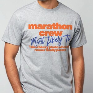 Könnte beinhalten: Graues T-Shirt mit dem Text "marathon crew" in orangefarbenen Blockbuchstaben über "Most Likely To" in blauer Schrift. Darunter steht der Satz "Take a blurry photo when the runner finally passes".