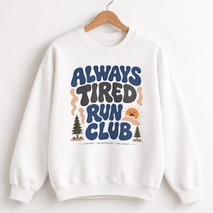May include: A white crewneck sweatshirt featuring a graphic with the text ALWAYS TIRED RUN CLUB in blue and dark gray wavy lettering. The design includes two pine trees, a sleepy orange sun, and the text SLOW PACE • LOW EXPECTATIONS • HIGH CAFFEINE.