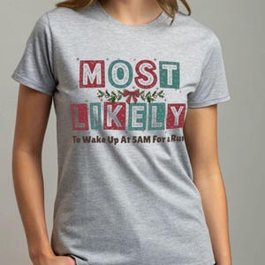 Könnte beinhalten: Graues T-Shirt mit dem Text "MOST LIKELY To Wake Up At 5AM For a Run" in roten und türkisfarbenen Blockbuchstaben. Der Text ist mit einer Schleife und Stechpalmenzweigen verziert, was auf ein Feiertags-Thema hindeutet.