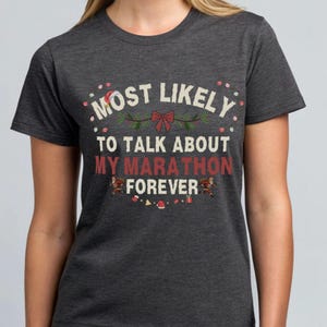 May include: Dark gray t-shirt with the text "MOST LIKELY TO TALK ABOUT MY MARATHON FOREVER" in white and red. The design includes festive accents like a bow, sprigs, and small ornaments, suggesting a holiday theme.