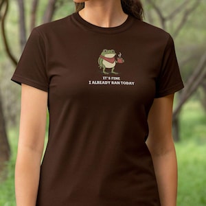 May include: A brown t-shirt featuring a cartoon frog wearing a red scarf and holding a coffee cup. The text "IT'S FINE I ALREADY RAN TODAY" is printed below the frog. The shirt is made of a soft material.