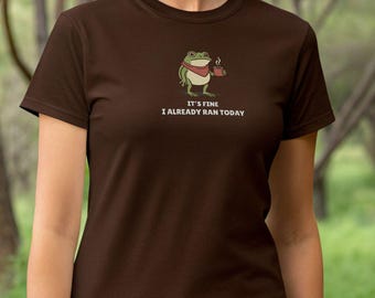 Pode incluir: Camiseta marrom com um sapo de desenho animado usando um cachecol vermelho e segurando uma xícara de café. O texto "IT'S FINE I ALREADY RAN TODAY" está impresso abaixo do sapo. A camiseta é feita de um material macio.