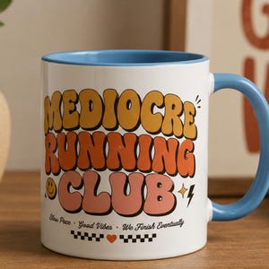 May include: A white ceramic mug with a light blue interior and handle. The front features the text MEDIOCRE RUNNING CLUB in large, wavy, multi-colored letters, along with a smiley face, lightning bolt, and the text Slow Pace, Good Vibes, We Finish Eventually.