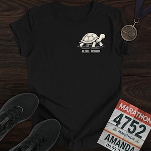 Puede incluir: Camiseta negra con una tortuga de dibujos animados y el texto "NO PACE NO PRESSURE". También se ven una medalla de maratón y zapatillas para correr. También se ve un dorsal de maratón con el nombre "AMANDA".