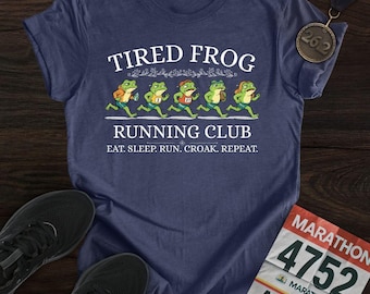 以下が含まれることがあります： 「TIRED FROG RUNNING CLUB」の文字と走るカエルのグラフィックがプリントされたネイビーブルーのTシャツ。「EAT. SLEEP. RUN. CROAK. REPEAT.」のフレーズも含まれています。マラソンメダルとゼッケンも表示されています。