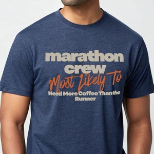 Könnte beinhalten: Marineblaues T-Shirt mit dem Text "marathon crew Most Likely To Need More Coffee Than the Runner" in Weiß und Orange. Das Shirt hat kurze Ärmel und einen Rundhalsausschnitt.