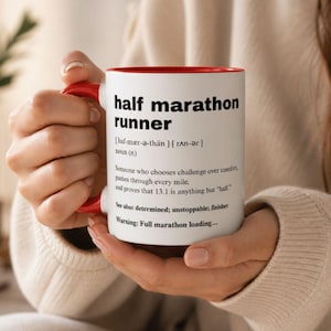 May include: White ceramic mug with a red interior and handle, featuring the text "half marathon runner" and a definition. The definition includes the phrase "pushes through every mile." The mug is held by a person.
