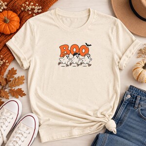 May include: A cream-colored short-sleeve t-shirt with a speckled texture. The center graphic features the orange text BOO with a small black bat above it, and three white ghost characters wearing hats and sneakers running in a line.