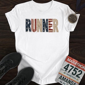 May include: White t-shirt with the word "RUNNER" in a patriotic design. The shirt is accompanied by a marathon medal, running shoes, and a race bib with the name "AMANDA" and the number "4752".
