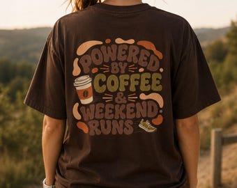 May include: A dark brown short-sleeve t-shirt featuring a graphic on the back with the text "POWERED BY COFFEE & WEEKEND RUNS". The design includes a coffee cup illustration, a running shoe, and abstract shapes in shades of tan, orange, and green.