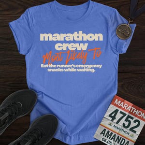 May include: A light blue t-shirt with the text "marathon crew Most Likely To Eat the runner's emergency snacks while waiting." A marathon medal and running shoes are also visible.