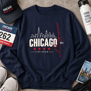 May include: A navy blue crewneck sweatshirt featuring a white Chicago skyline graphic, the text CHICAGO FINISHER, four red six-pointed stars, and a red vertical line map outline. The design includes the number 26.2 in white near the skyline.