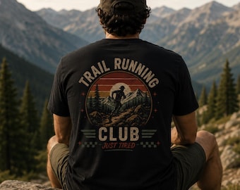 May include: A black t-shirt featuring a circular graphic on the back with a sunset, mountain range, and a runner. The text reads "TRAIL RUNNING CLUB" in an arc above and "JUST TIRED" below, accompanied by small star and checkered flag icons.