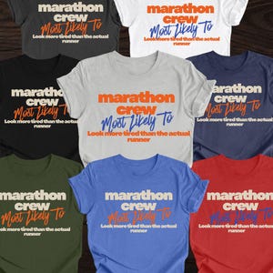 Könnte beinhalten: Eine Auswahl an T-Shirts in verschiedenen Farben, darunter Schwarz, Weiß und Blau. Jedes Shirt zeigt den Text "Marathon Crew Most Likely To Look more tired than the actual runner" in einem auffälligen Design. Die Shirts sind wahrscheinlich für Marathon-Unterstützer.