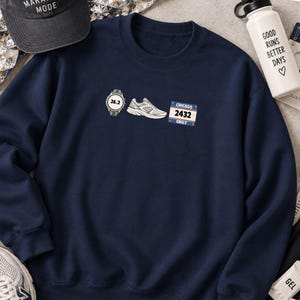 Pode incluir: Uma camisola de gola redonda azul-marinho com uma impressão gráfica de um relógio desportivo digital a mostrar 26.2, um sapato de corrida cinzento e um dorsal da maratona de Chicago com o número 2432 e o nome Emily em azul, branco e vermelho.