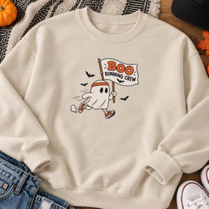 May include: A beige crewneck sweatshirt featuring a graphic of a cartoon ghost wearing a headband and trainers, running while holding a flag that reads "BOO RUNNING CREW" in orange and black text, surrounded by small black flying bats.