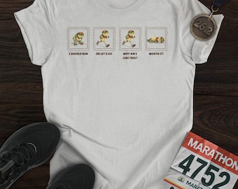 May include: A white t-shirt featuring a frog design in a stamp-like style, illustrating marathon stages. Text on the shirt includes "I SHOULD RUN", "OK LET'S GO", "WHY AM I LIKE THIS?", and "WORTH IT!". A marathon medal and race bib are also present in the image.