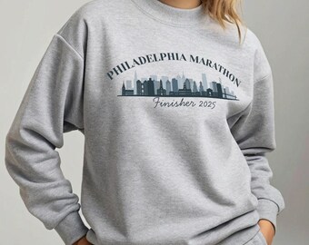 Puede incluir: Una sudadera gris con el texto "PHILADELPHIA MARATHON Finisher 2025" arqueado sobre un gráfico de horizonte azul oscuro. La sudadera tiene cuello redondo y mangas largas. La persona lleva un pantalón de chándal gris a juego.