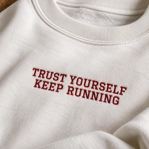 May include: An off-white crewneck sweatshirt featuring the embroidered text TRUST YOURSELF KEEP RUNNING in a bold, dark red font centered on the chest. The garment has a ribbed collar and a soft, textured fabric construction.
