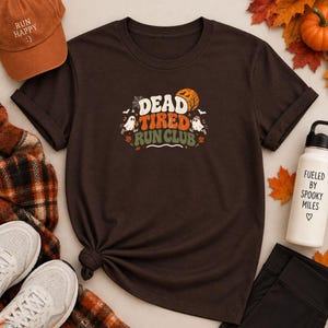 Puede incluir: Una camiseta de manga corta de color marrón oscuro con un gráfico que incluye el texto "DEAD TIRED RUN CLUB" en blanco y naranja. El diseño presenta dos pequeños fantasmas blancos, una telaraña, murciélagos y una luna naranja sobre fondo marrón.