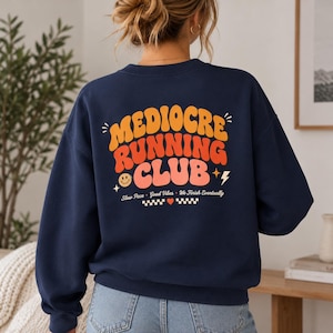 Pode incluir: Uma camisola azul-marinho de gola redonda com um gráfico nas costas com o texto "MEDIOCRE RUNNING CLUB" em letras onduladas laranja, vermelhas e cor-de-rosa. Abaixo do texto estão um smiley, um raio e as palavras "Slow Pace • Good Vibes • We Finish Eventually" com um pequeno coração vermelho e padrões de xadrez.
