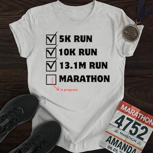 May include: A light gray t-shirt with a checklist design for running distances: 5K, 10K, 13.1M, and Marathon. The shirt includes a marathon medal and a race bib. Black running shoes are at the bottom.