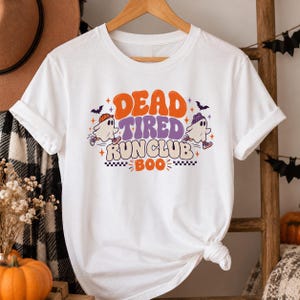 May include: A white short-sleeve t-shirt featuring a graphic with the text DEAD TIRED RUN CLUB BOO. The design includes two cartoon ghosts wearing beanies and sneakers, surrounded by small stars and bats in orange and purple colors.