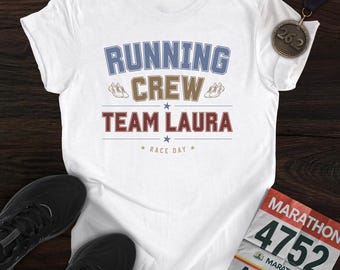 May include: A white short-sleeved t-shirt featuring the text RUNNING CREW TEAM LAURA RACE DAY in blue, brown, and red, with small shoe icons and stars. A bronze 26.2 medal and a marathon race bib numbered 4752 for AMANDA are placed nearby.