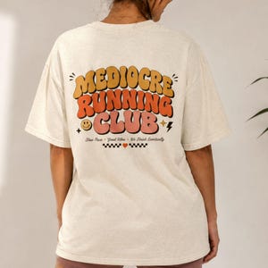May include: A cream-colored short-sleeve t-shirt featuring a graphic on the back with the text "MEDIOCRE RUNNING CLUB" in large, wavy, orange and pink letters. Below the text, it reads "Slow Pace • Good Vibes • We Finish Eventually" with a checkerboard pattern and a small heart.