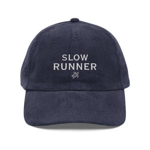 May include: Navy blue corduroy baseball cap with the text "SLOW RUNNER" embroidered in white, along with a small turtle graphic. The cap has a curved brim and a fabric strap closure.
