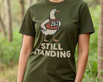 May include: An olive green t-shirt with a cartoon duck wearing a running bib marked "26.2". Below the duck is the text "STILL STANDING" in a bold, off-white font. The t-shirt has a crew neck and is made of a soft material.