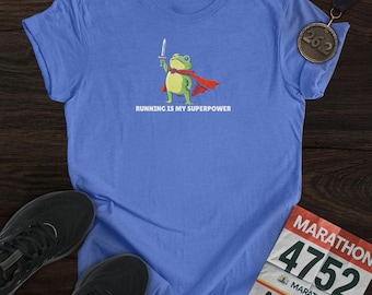 May include: A light blue t-shirt featuring a cartoon frog superhero with a sword and cape, with the text "RUNNING IS MY SUPERPOWER". A marathon medal and running shoes are also visible in the image.