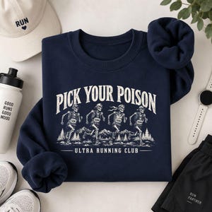May include: A navy blue crewneck sweatshirt featuring a white graphic of four skeletons running in a mountain landscape. The text reads PICK YOUR POISON and ULTRA RUNNING CLUB, with the skeletons wearing race bibs labeled 50K, 50MI, 100K, and 100MI.
