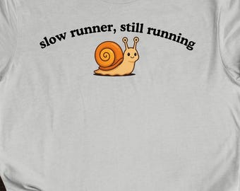 以下が含まれることがあります： 「slow runner, still running」の文字と、茶色の殻と笑顔の顔を持つカタツムリの漫画が描かれた、薄い灰色のTシャツ。 カタツムリは薄茶色で走っています。