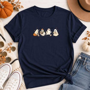 May include: A navy blue short-sleeve t-shirt featuring a central graphic of four white ghosts. The ghosts are depicted in various activities, including sitting on a pumpkin, holding a stack of books, running, and reading a small book.