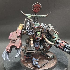 Op de afbeelding: Gedetailleerde miniatuur van een Ork-oorlogsmachine, met een groenhuidige Ork-piloot. Het model is geschilderd in tinten rood, groen, bruin en metaal, met een grote klauw, een multi-loop geweer en een dambordpatroon.
